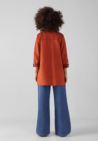 Abrigo de ante terracota con corte recto y mangas enrolladas, combinado con pantalones de mezclilla azul de pierna ancha. Textura suave y diseño sencillo.