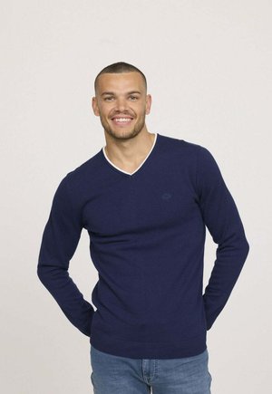 Pull à col en V bleu marine en tissu maille lisse, avec un bord blanc au niveau de l'encolure et des poignets côtelés. Porté avec un jean bleu clair.