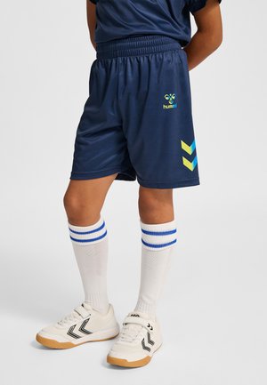 Barn iført marineblå sportsshorts med gule og blå chevronmønstre, hvide knæstrømper med blå striber og hvide sportssko.