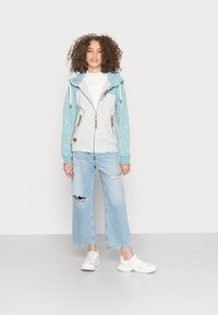 Camisola de capuz cinza claro com fecho, mangas impressas em turquesa e pormenores em madeira, combinada com jeans largos claros desgatados e ténis brancos.