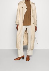 Trench-coat en cuir beige avec un col classique, un devant à double boutonnage et des poches latérales, associé à un pull marron et un pantalon crème.