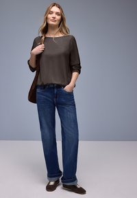 Blouse à manches longues gris foncé avec un large décolleté, associée à un jean bleu ample et des chaussures marron, présentée sur un fond neutre.
