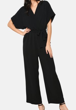 Femme portant une combinaison noire à manches courtes avec jambes larges et une ceinture en tissu nouée à la taille, debout une main sur la hanche.