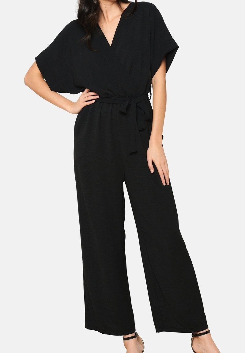 Femme portant une combinaison noire à manches courtes avec jambes larges et une ceinture en tissu nouée à la taille, debout une main sur la hanche.