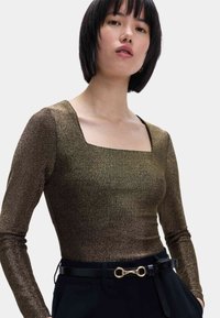 Top de manga larga en tejido metálico bronce con escote cuadrado. Combinado con un pantalón negro con cinturón que presenta un detalle de hardware dorado.