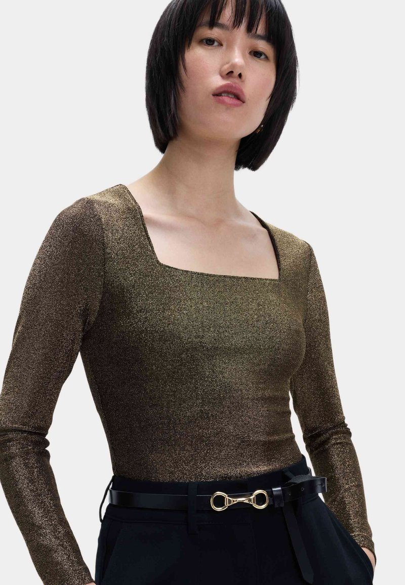 Top de manga larga en tejido metálico bronce con escote cuadrado. Combinado con un pantalón negro con cinturón que presenta un detalle de hardware dorado.