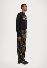 Black sweatshirt with gold "VERSACE JEAN COUTURE" text, paired with black pants featuring a gold chain pattern, worn with black sneakers.