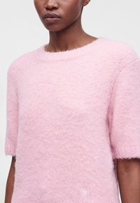 Άτομο που φοράει ένα απαλό, χνουδωτό, ανοιχτό ροζ πουλόβερ με κοντά μανίκια και στρογγυλή λαιμόκοψη σε ουδέτερο φόντο.