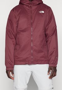 Mężczyzna ubrany w bordową kurtkę z kapturem The North Face na zamek błyskawiczny z elastycznymi mankietami oraz białe spodnie z kieszenią na udzie zapinaną na zamek.