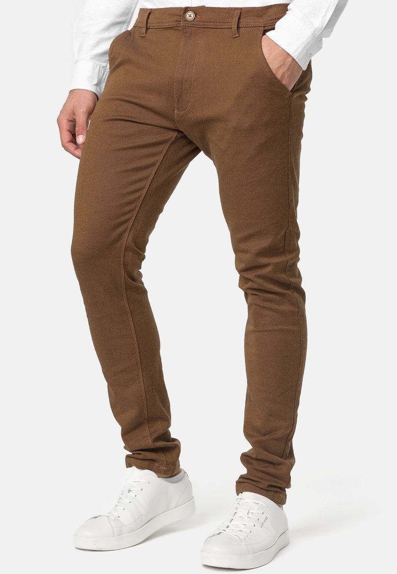 Pantalon marron slim porté avec une chemise blanche à manches longues et des baskets blanches, une main dans la poche, sur fond blanc uni.