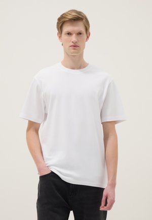 Joven con cabello corto castaño claro, vestido con una camiseta blanca de manga corta lisa y pantalones negros, de pie con una mano en el bolsillo.