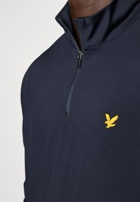 Marineblauer Quarter-Zip-Pullover aus glattem Stoff, mit einem gelben Adler-Logo auf der Brust, das das schlichte Design betont.