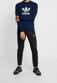 Námořnická mikina s bílým logem Adidas, doplněná černými tepláky s malým emblémem Adidas. Model má na sobě černé sportovní boty.