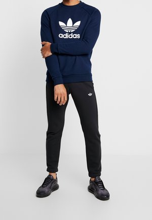 Granatowa bluza z kapturem z białym logo Adidas, zestawiona z czarnymi dresami z małym emblematem Adidas. Model ma na nogach czarne buty sportowe.