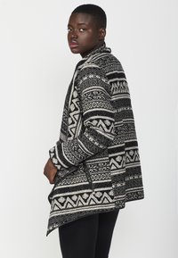 Schwarz-weiße, gemusterte Jacke mit legerem Schnitt, die geometrische Designs aufweist, einen Kragen und lange Ärmel hat. Sie besteht aus einem strukturierten Stoff.