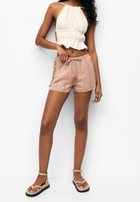 Jasnobeżowy krótki top z marszczonym dekoltem, zestawiony z różowymi szortami w wzory z złotymi akcentami i sznurkiem do regulacji. Brązowe sandały.
