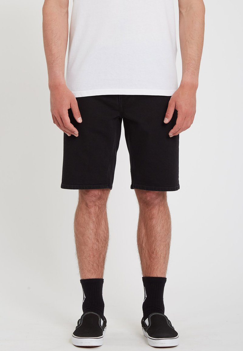 Shorts en denim noir avec une coupe droite, longueur genoux, et un design à cinq poches. Portés avec des chaussettes noires et des chaussures à enfiler.