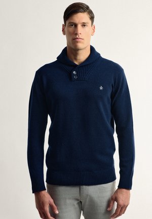 Suéter de punto azul marino con cuello alto, que presenta tres botones, puños y dobladillo acanalados. Bordado sutil en el pecho. Textura suave.