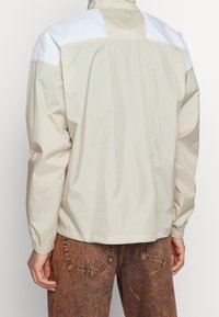 Beige vindjacka med en vit axelpanel, har en zippered hals och elastiska mudd. Bärs över bruna strukturerade byxor.