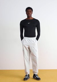 Tricou sport lung de culoare neagră, cu logo-ul alb Nike, asociat cu pantaloni cargo de culoare gri deschis, cu croială lejeră, și adidași sport negri.