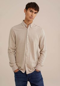 Camisa de manga larga beige con cuello abotonado, que presenta un pequeño logo en el pecho, combinada con jeans azules. Textura lisa y uniforme.