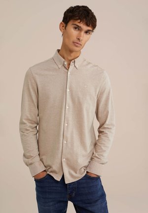 Beige long-sleeve shirt with a button-down collar, featuring a small logo on the chest, paired with blue jeans. Smooth, plain texture.