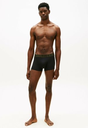 Junger schwarzer Mann, barfuß, trägt schwarze Boxer-Shorts mit einem dunkelgrünen Bund vor einem schlichten weißen Hintergrund.