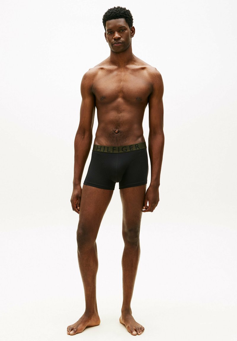 Young Black male standing barefoot in black boxer briefs with a dark green waistband on a plain white background.