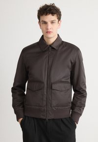 Veste aviateur marron avec une texture lisse, fermeture éclair à l'avant, col plat et deux grandes poches avant. Les poignets et la taille côtelés complètent le design.