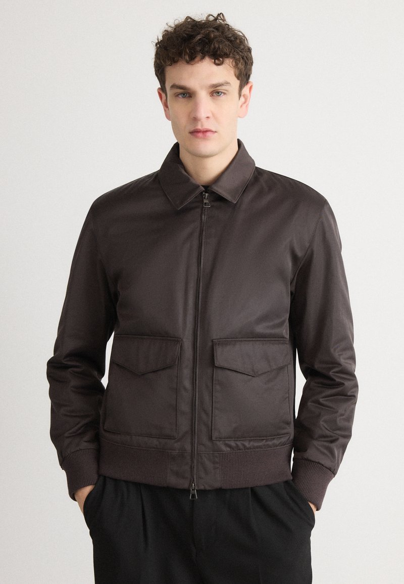 Veste aviateur marron avec une texture lisse, fermeture éclair à l'avant, col plat et deux grandes poches avant. Les poignets et la taille côtelés complètent le design.