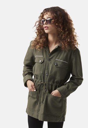Femme aux cheveux bouclés portant des lunettes de soleil foncées et une veste vert olive boutonnée avec taille à cordon et poches poitrine cloutées.