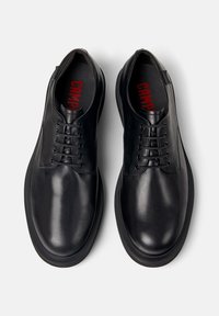 Pantofi din piele neagră cu șireturi și talpă groasă, văzuți dintr-o perspectivă de deasupra pe un fundal alb, cu branding roșu "CAMPER" în interior.