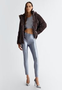 Giacca imbottita marrone scuro, abbinata a un top corto grigio chiaro e leggings grigi lucidi. Scarpe argentate con tacco alto completano l'outfit.