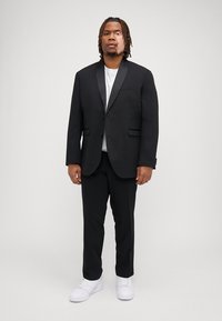 Black suit with a notch lapel and single button closure, paired with a white t-shirt and white sneakers. Model stands against a neutral background.