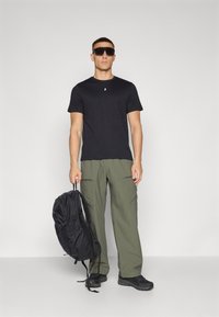 Sort kortærmet t-shirt, olivengrøn cargo-bukser med lynlåslommer, sort elegant rygsæk og sorte sko, præsenteret mod en ensfarvet baggrund.