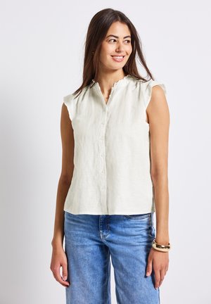 Blusa sin mangas de lino color beige claro con un cuello con volantes, cierre de botones en la parte delantera y un corte relajado. Combinada con unos jeans de mezclilla azul.