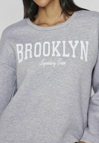 Sudadera gris con cuello redondo, que presenta letras blancas "BROOKLYN" y "Legendary Team" impresas en la parte frontal. Textura suave.