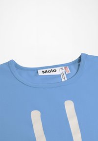 Modrá tričko s okrúhlym výstrihom, s bielymi vertikálnymi pruhmi a nápisom značky "Molo" našitým vo vnútri goliera, so zobrazenou veľkosťou 122/128 a štítkami s pokynmi na údržbu.