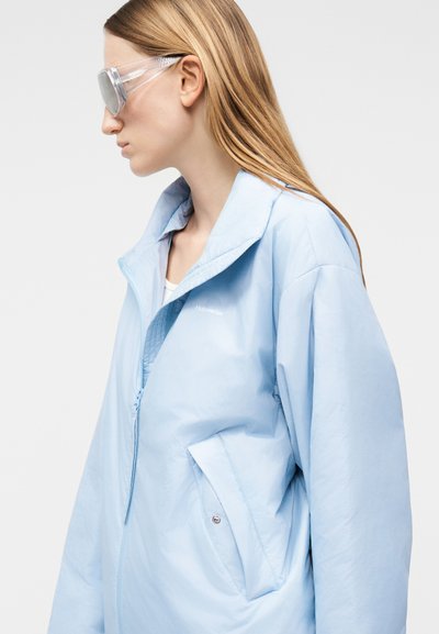 Γυναίκα με μακριά ξανθά μαλλιά φορά υπερμεγέθη ανοιχτό μπλε μπουφάν και διαφανή γυαλιά προστασίας, κοιτάζοντας προς τα αριστερά σε απλό λευκό φόντο.