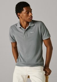 Sivá polokošeľa s textúrovanou látkou, zapínaním na tri gombíky a vyšívaným logom na ľavej strane hrude, v kombinácii so svetlými nohavicami.