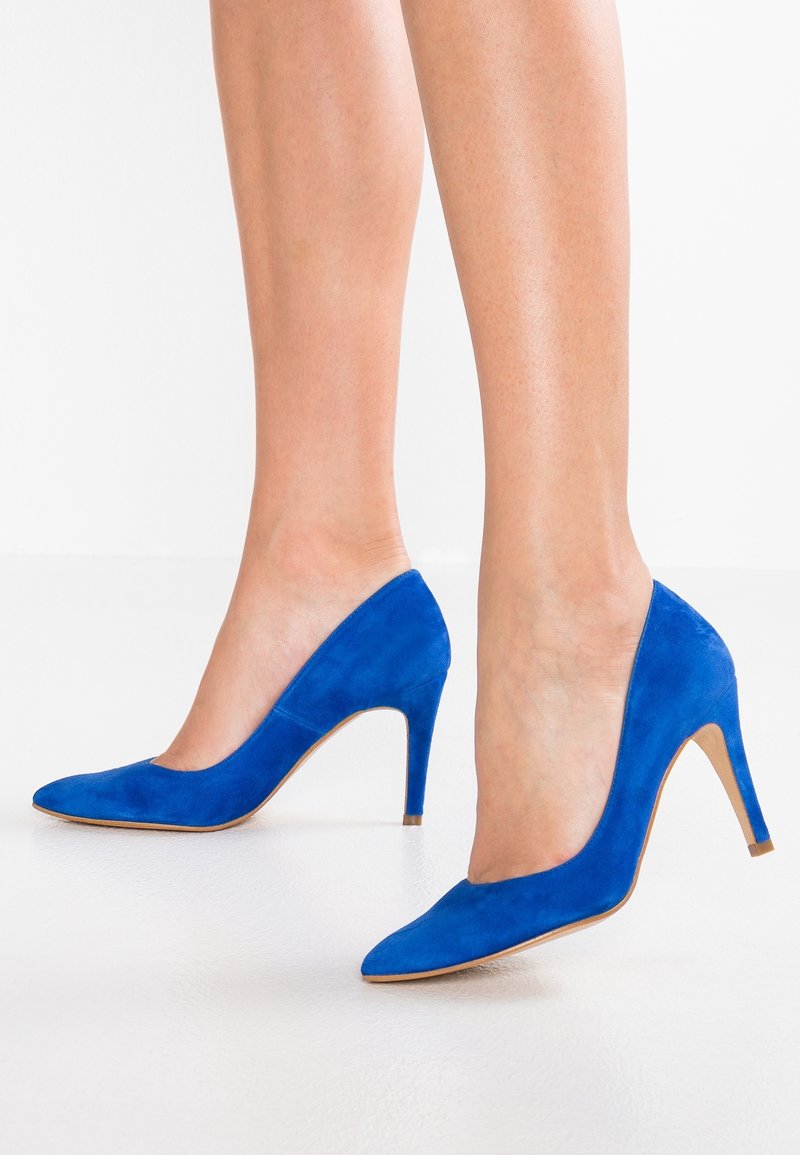 Jambes de femme portant des escarpins en daim bleu vif à bout pointu avec des talons aiguilles fins sur une surface blanche.