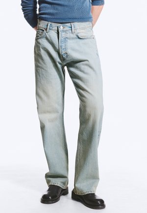 Personne portant un jean bleu clair, des chaussures noires et une chemise bleue à manches longues, debout les mains derrière le dos.