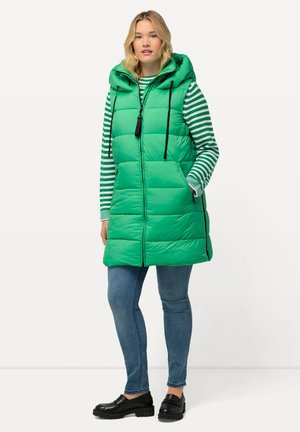Gilet imbottito verde con cappuccio, caratterizzato da una chiusura con zip, lacci neri e tasche laterali; indossato sopra una maglietta a righe orizzontali e jeans blu.