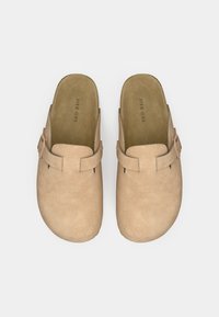 Beige színű velúr klumpák zárt orral és nyitott hátsó résszel, mindkét cipőn egy-egy díszes csattal és textúrázott talpbéléssel.