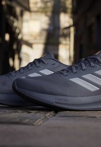 Chaussures de course Adidas grises avec des bandes blanches sur une rue pavée, éclairées par la lumière du soleil avec un arrière-plan urbain flou.
