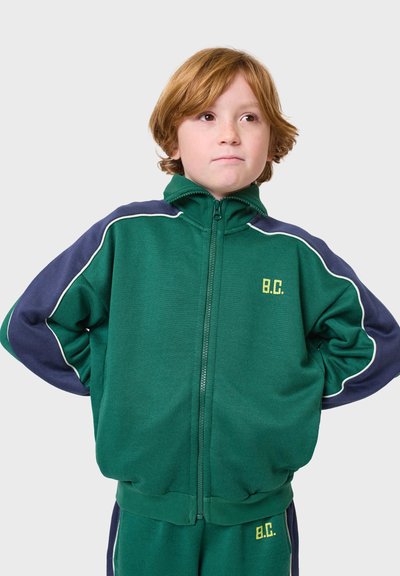 Zelená traková bunda na zips s námorníckom modrými rukávmi a bielym lemovaním. Má okrúhly golier a vyšitú iniciálu "B.C." v žltej farbe. Mäkká látka.