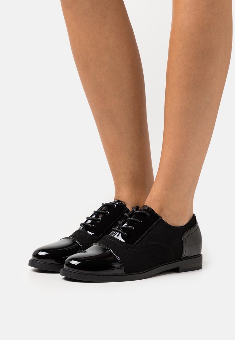 Chaussures Oxford noires à lacets avec bouts en cuir brillant et côtés en daim lisse portées pieds nus sur fond blanc.