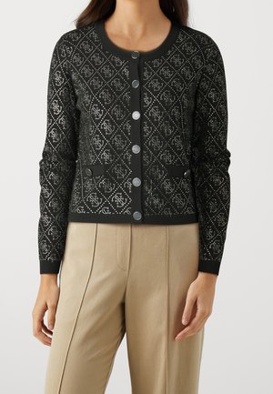 Femme portant un cardigan noir boutonné avec un motif en losanges et un pantalon beige taille haute sur fond uni.