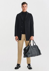 Czarny wełniany płaszcz na czarnym golfie, beżowe spodnie oraz czarna skórzana torba podróżna z podwójnymi uchwytami. Całość dopełniają eleganckie mokasyny.