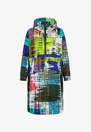 Femme portant une longue veste à motifs abstraits colorés, un jean bleu et des baskets violettes, debout devant un fond blanc uni.